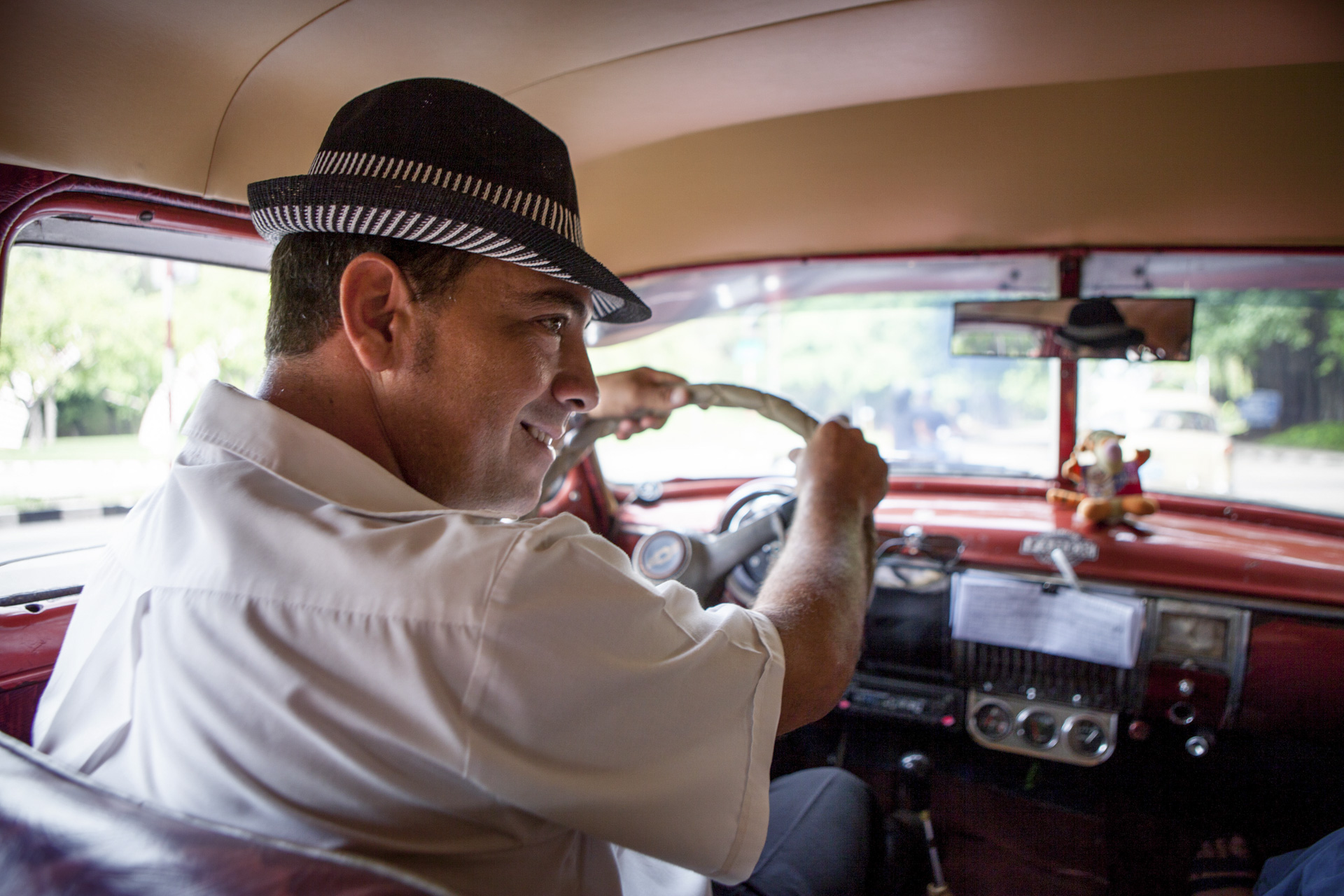 Young male taxidriver wearing a hat, inside of his american taxi, car, in Havana Cuba.
