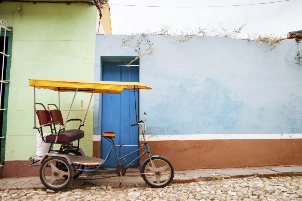 En tom cykeltaxi, parkeret foran en grøn og en blå væg på en brostensbelagt gade i byen Trinidad på Cuba. Empty taxi bike in Trinidad, Cuba.